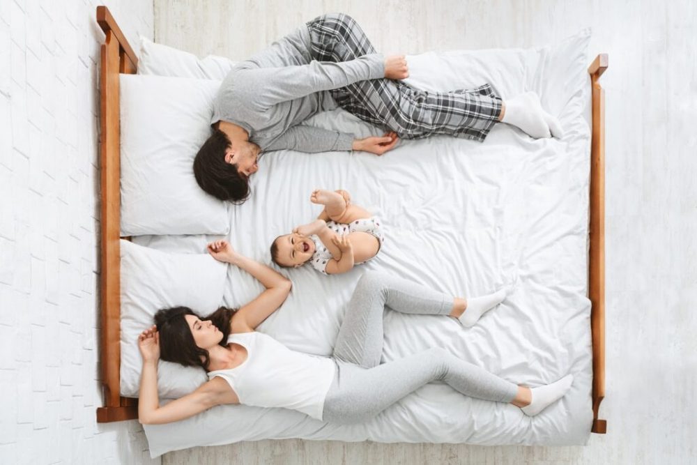 Family on a bed with sleepy parents pushed aside and a happy baby in the middle