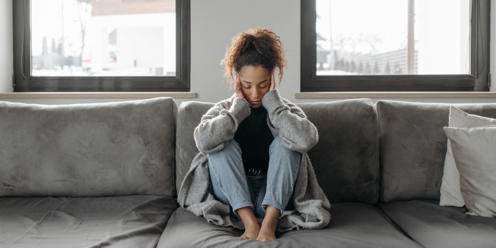 A woman is sitting on a gray couch. She is having a headache.